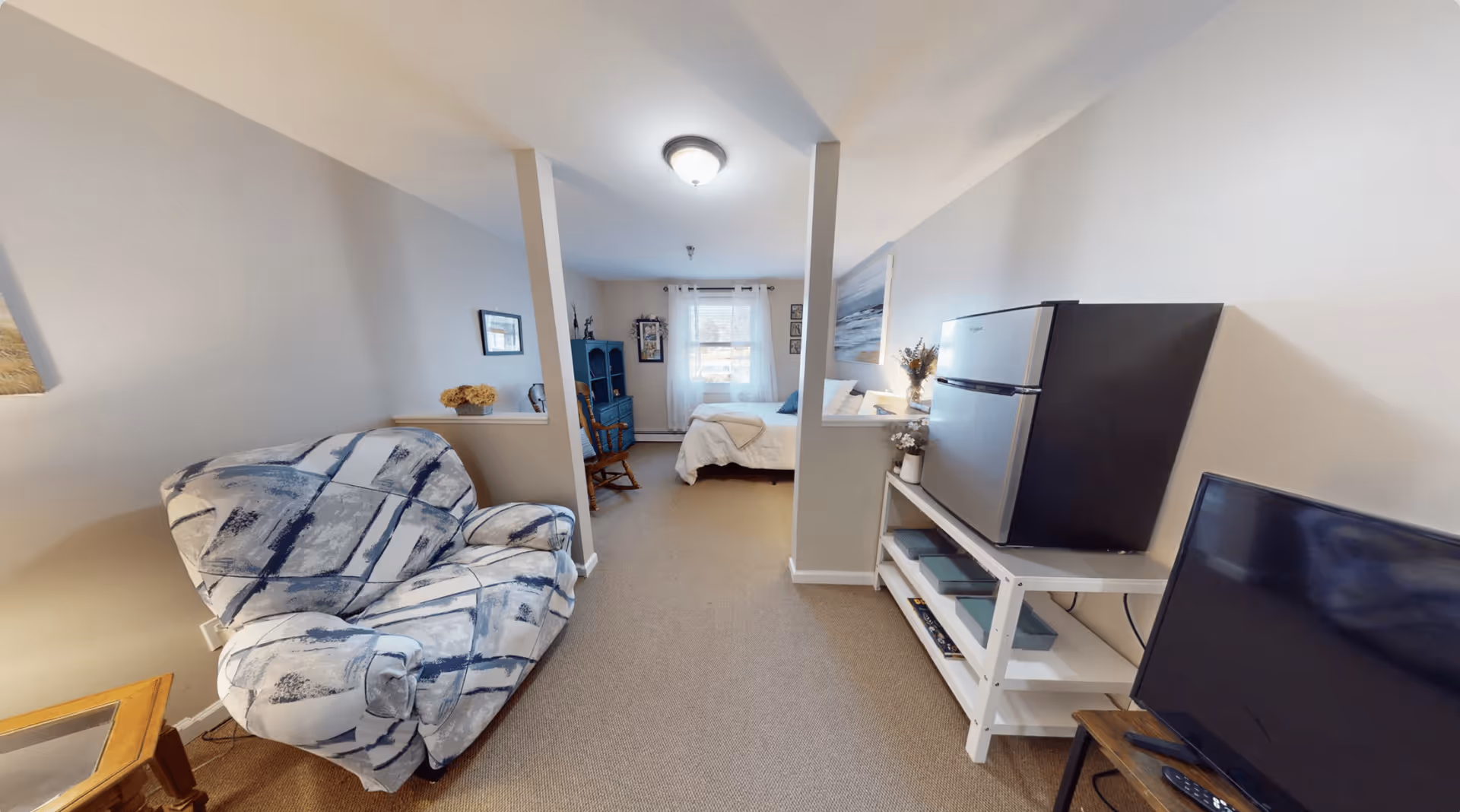 Interior view of a senior living facility room featuring a patterned armchair on the left, a small refrigerator on a white shelf on the right, a television on a stand, and a bed with white bedding in the background near a window with sheer curtains.