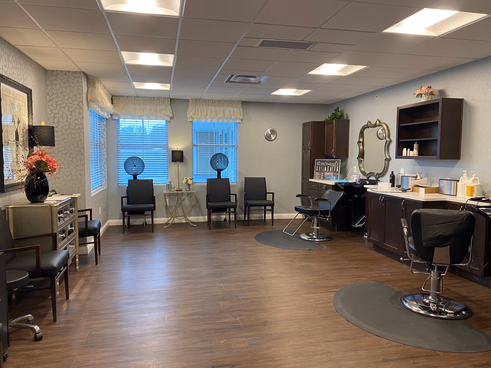 A spacious salon room with wooden flooring and light gray walls. The room features two black salon chairs with footrests on circular mats, a large ornate mirror on the wall, and dark wood cabinetry with various hair care products. There are three black chairs along the far wall beneath two windows with white blinds and valances. Two black hair dryers are positioned near the chairs. A small table with a lamp and flowers is placed between the windows. Additional seating and a decorative chest with a vase of flowers are on the left side of the room.