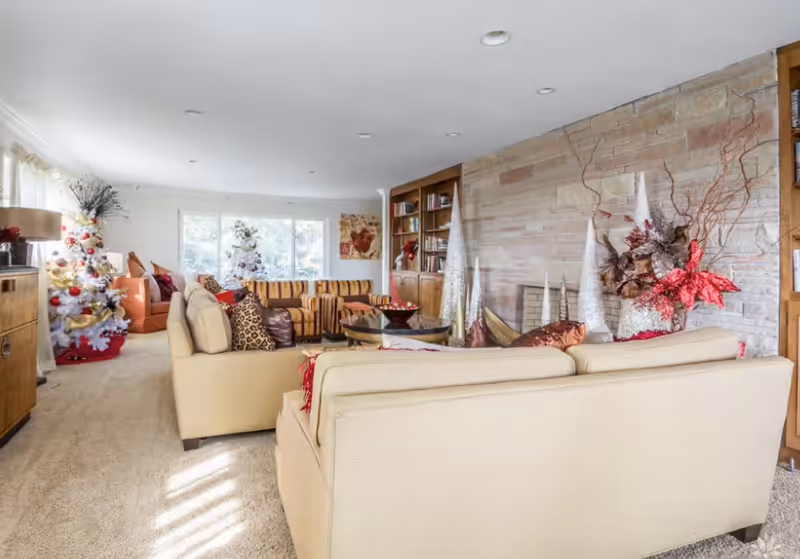 A spacious living room decorated for the holidays with two white Christmas trees adorned with gold and red ornaments, a beige sectional sofa with various pillows, a striped sofa, a wooden bookshelf, and festive red and gold floral arrangements on a stone accent wall.