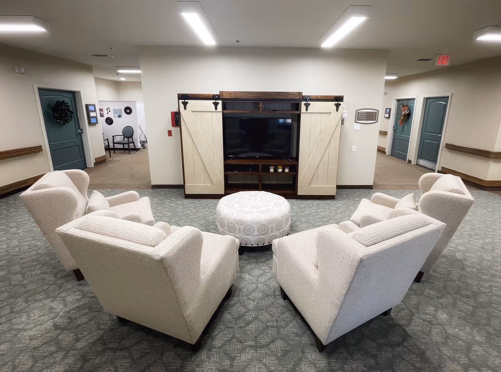 Small lounge area with four beige upholstered armchairs and a patterned ottoman facing a TV in a wooden cabinet in a senior living facility hallway.
