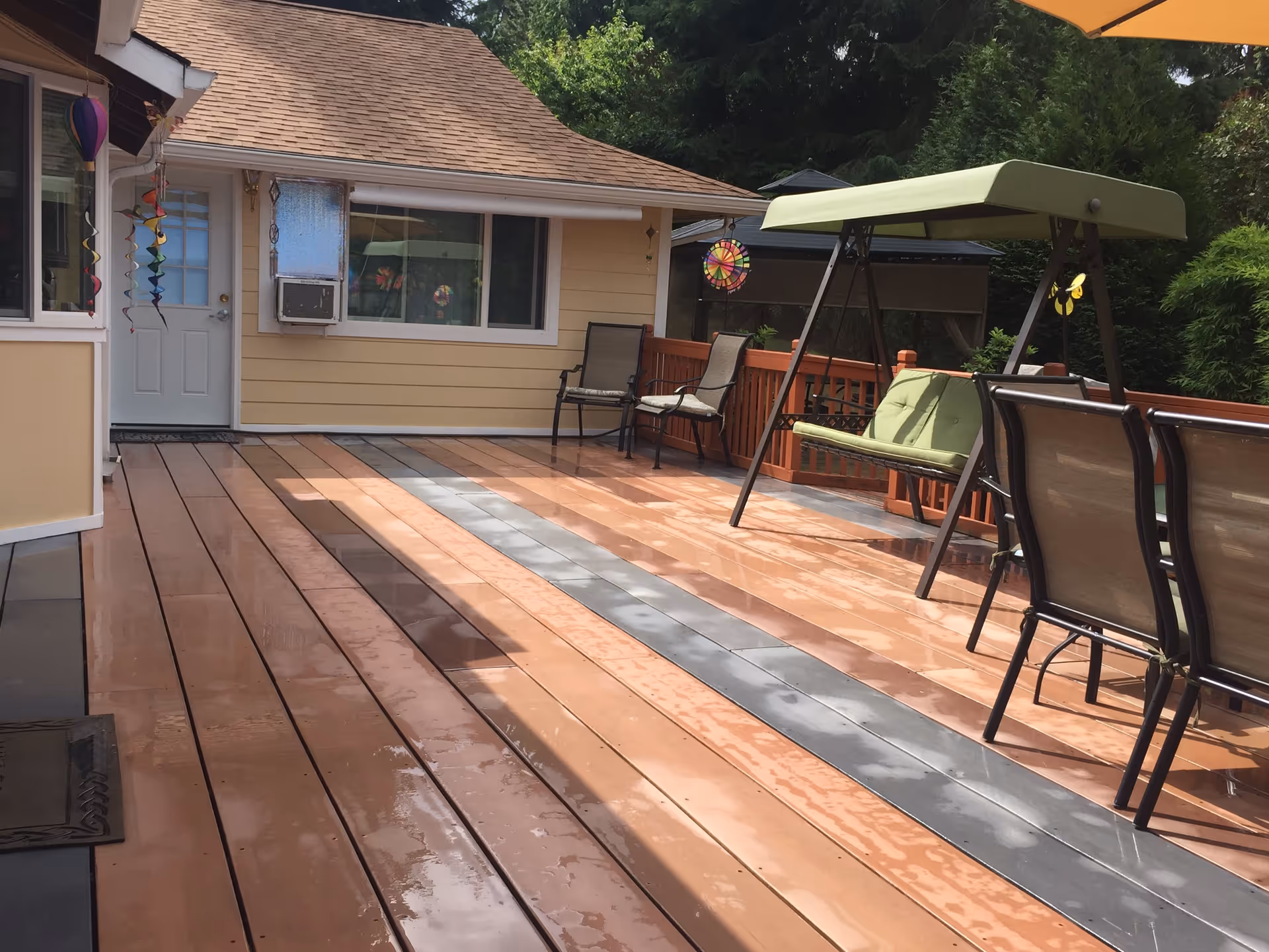 Outdoor wooden deck area with wet floor, featuring a green cushioned swing, several chairs, and a small table. The deck is attached to a beige building with a white door and window, decorated with colorful hanging ornaments. Trees and greenery are visible in the background.