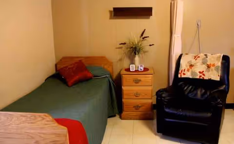 A small senior living bedroom with a bed, nightstand, chair, and decorative plant arrangement.