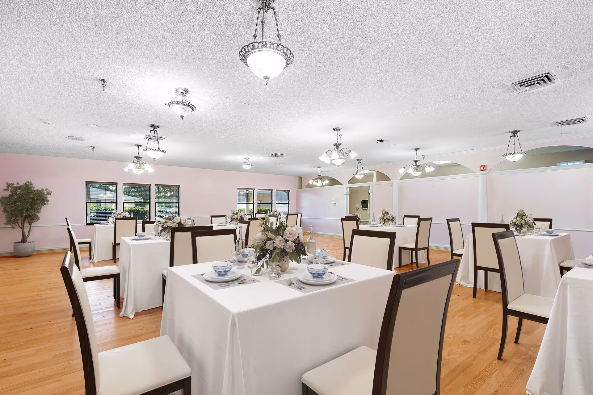 Bright dining room with multiple tables dressed in white tablecloths, floral centerpieces, and chairs on a hardwood floor.