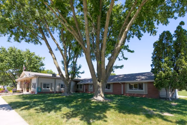 Single-story brick and siding building with large trees and green grass in front under a clear blue sky.