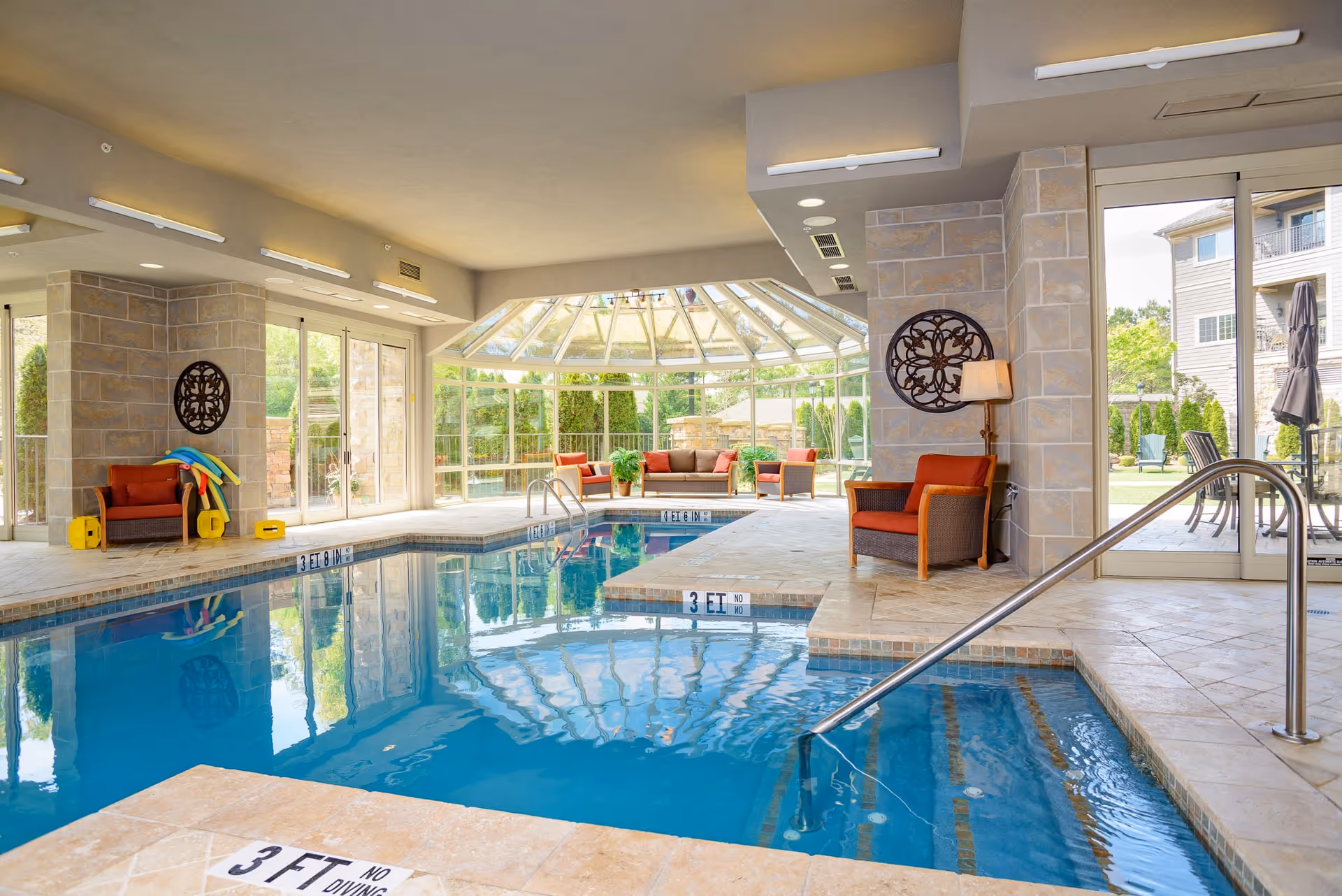 Indoor swimming pool area with clear blue water, surrounded by beige tiled flooring and stone pillars. There are comfortable wicker chairs with orange cushions and pool noodles stacked on one chair. Large windows and glass doors allow natural light to fill the space, and outside greenery is visible. A metal handrail leads into the pool, and a seating area with sofas and plants is visible in a sunroom extension.