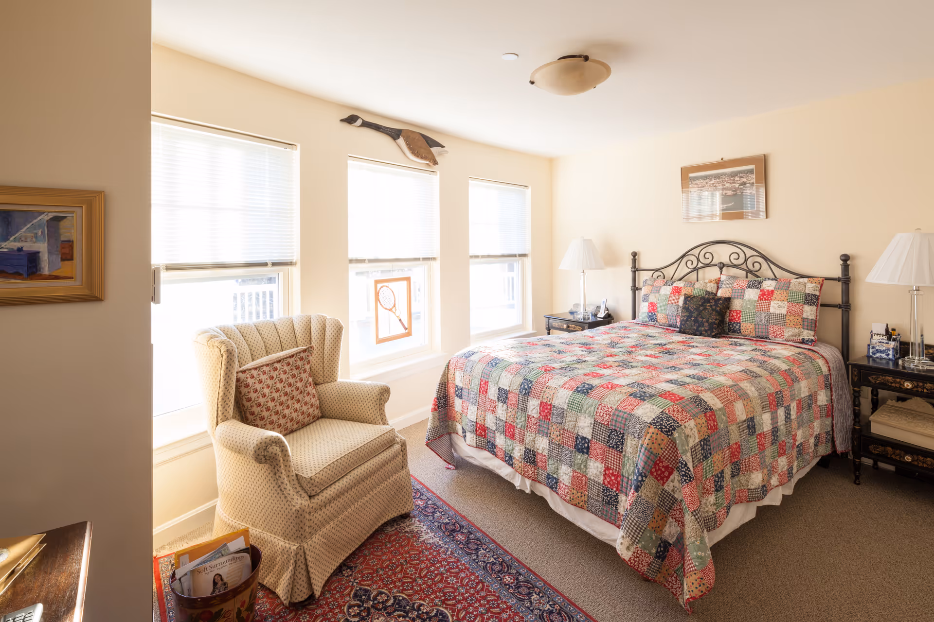 A bright and cozy bedroom with a large bed covered in a colorful patchwork quilt. The room has three windows with white blinds, a comfortable upholstered armchair with a floral cushion, two bedside tables with lamps, and framed artwork on the walls. A red patterned rug partially covers the carpeted floor.