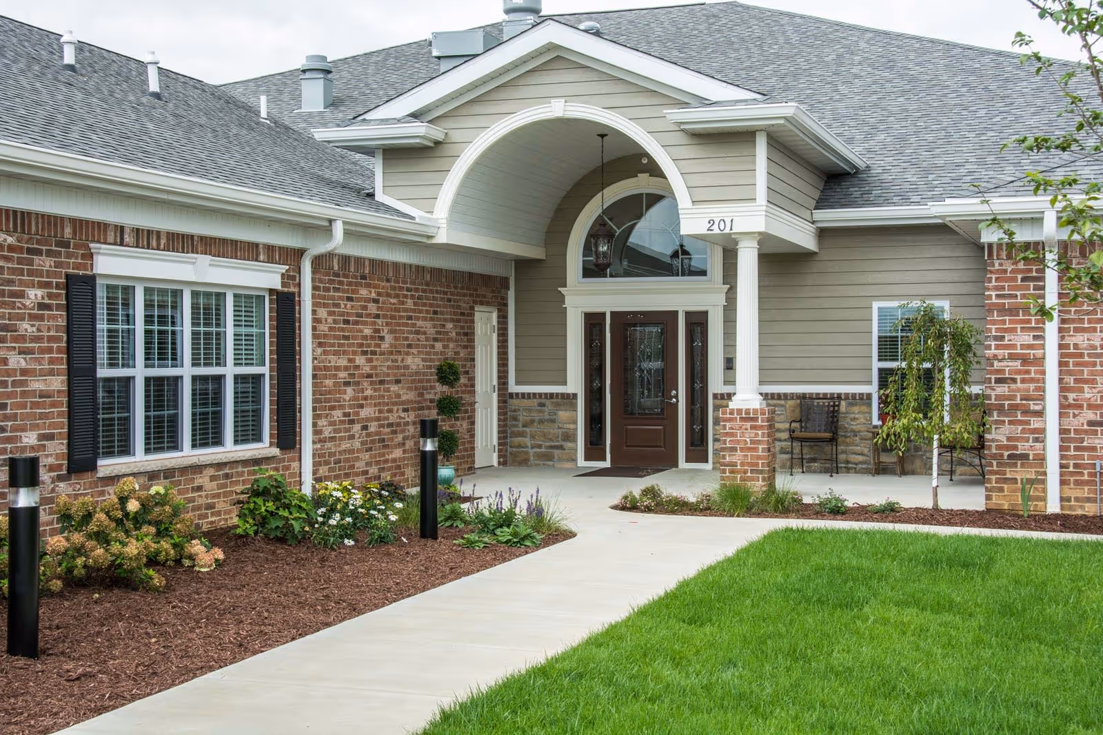 Front entrance of a brick and siding senior living building with a covered portico and landscaped walkway.