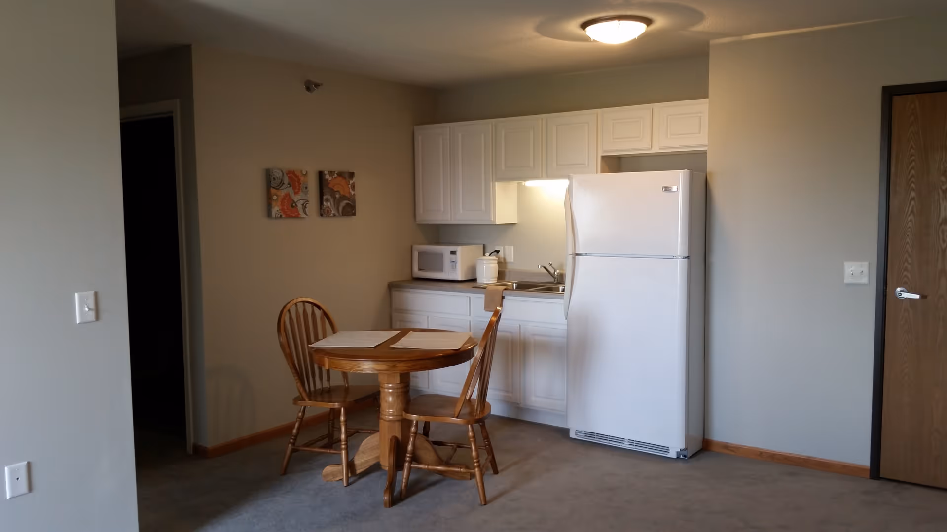 A small kitchen area with white cabinets, a white refrigerator, a microwave, and a sink. In front of the kitchen is a round wooden table with two wooden chairs. The walls are painted beige, and there are two small decorative paintings on the wall near the kitchen.