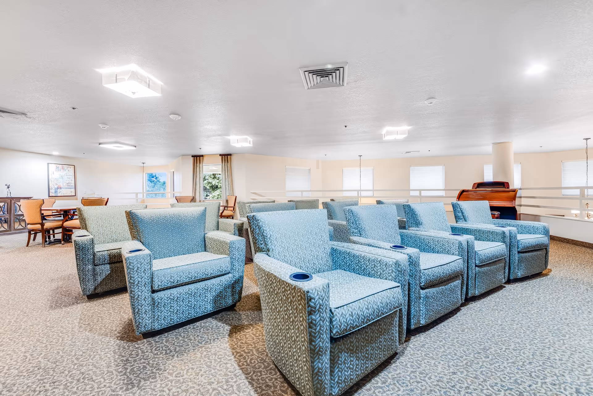 A spacious, well-lit common area with multiple light blue upholstered armchairs arranged in rows, each featuring a built-in cup holder. In the background, there are wooden tables with chairs and a piano against the wall. The room has beige walls, carpeted floors, and several windows letting in natural light.