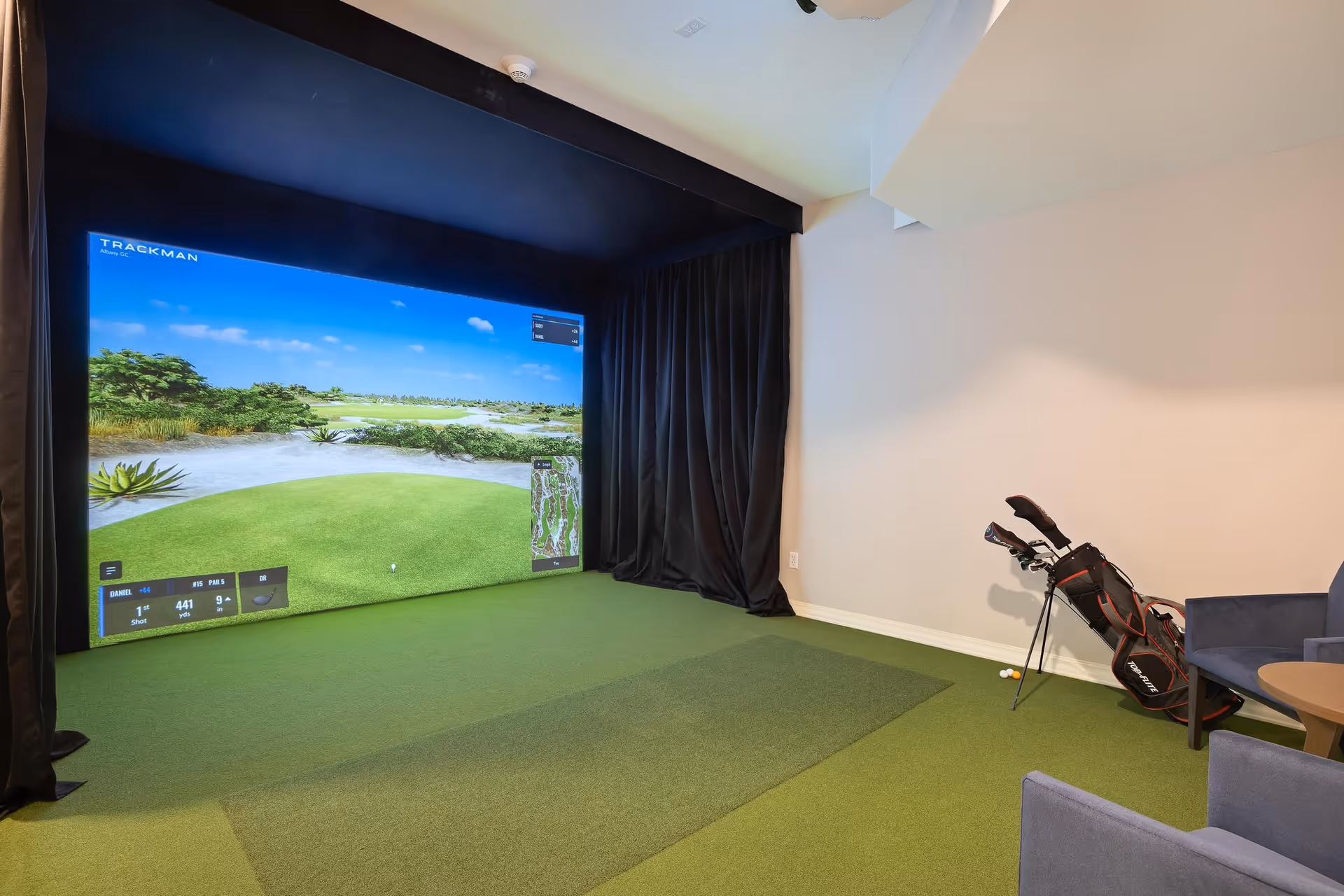 Indoor golf simulator room with a large screen displaying a golf course, green carpet flooring, a golf bag with clubs, and two blue chairs with a small round table.