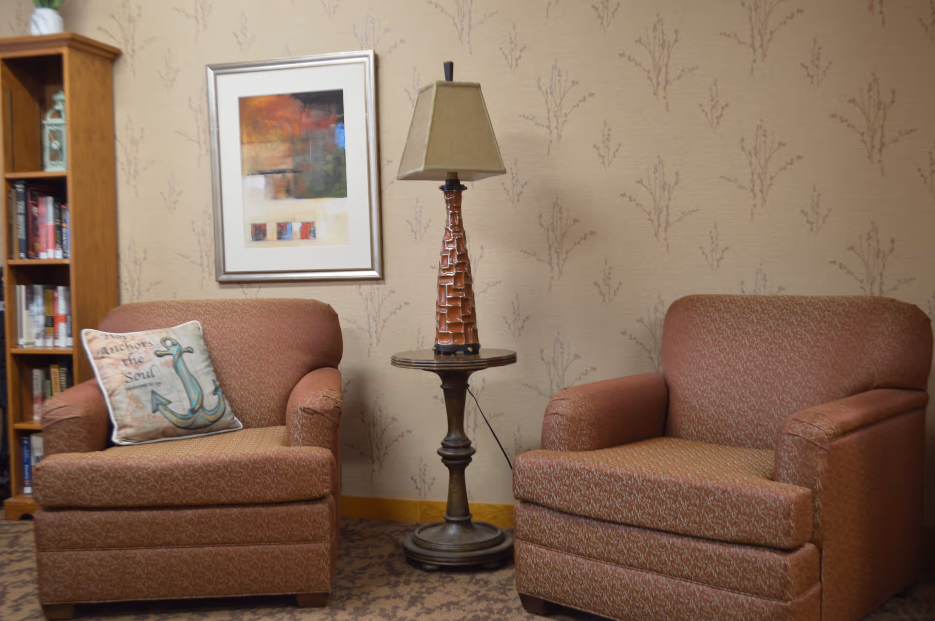 A cozy sitting area with two brown upholstered armchairs separated by a round wooden side table with a decorative lamp on it. One armchair has a pillow with an anchor design and the words 'Anchors the Soul'. Behind the chairs is a wall with subtle patterned wallpaper, a framed abstract painting, and a wooden bookshelf filled with books.