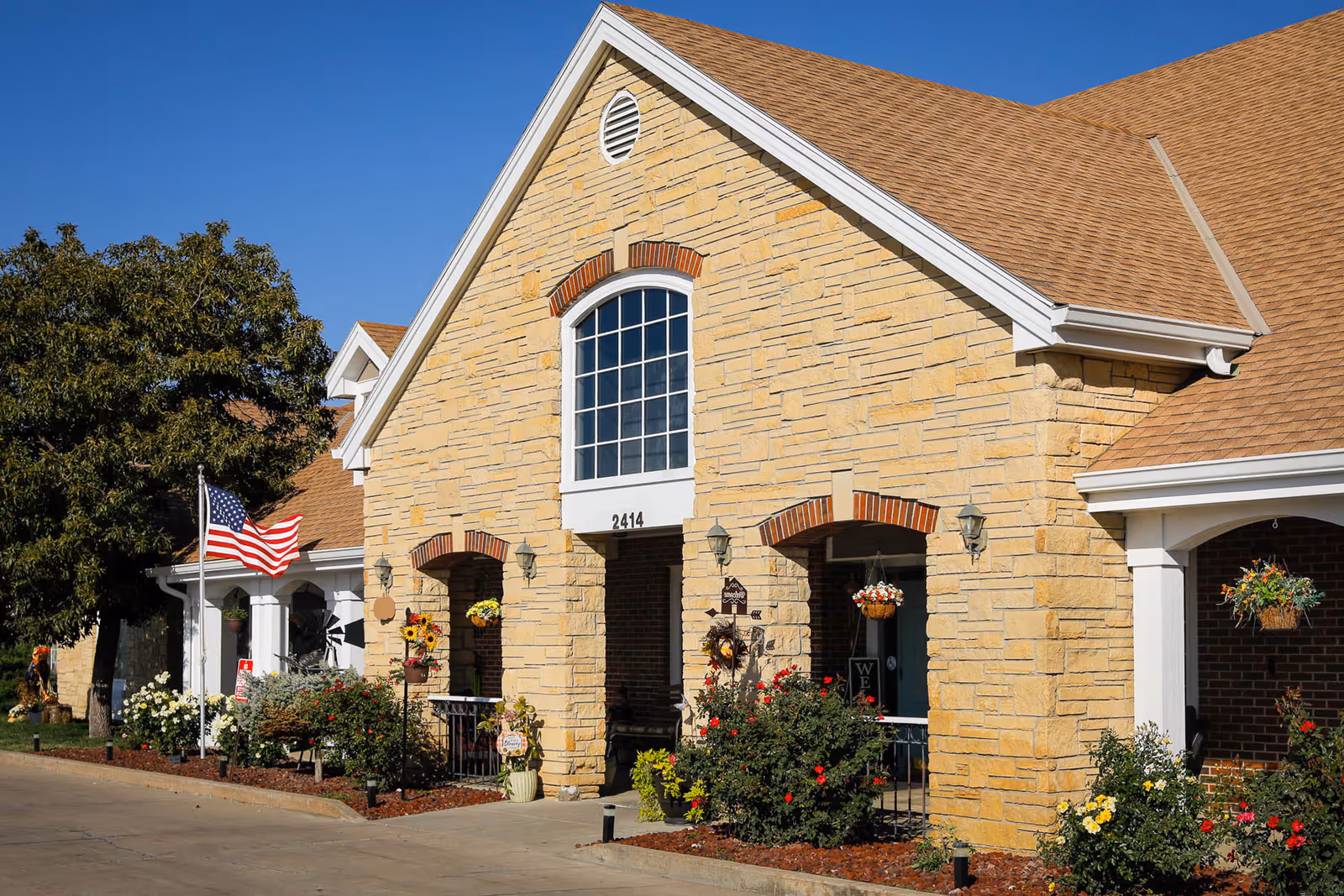 Front exterior of a stone-faced assisted living building with an arched window, American flag, and landscaped flower beds.