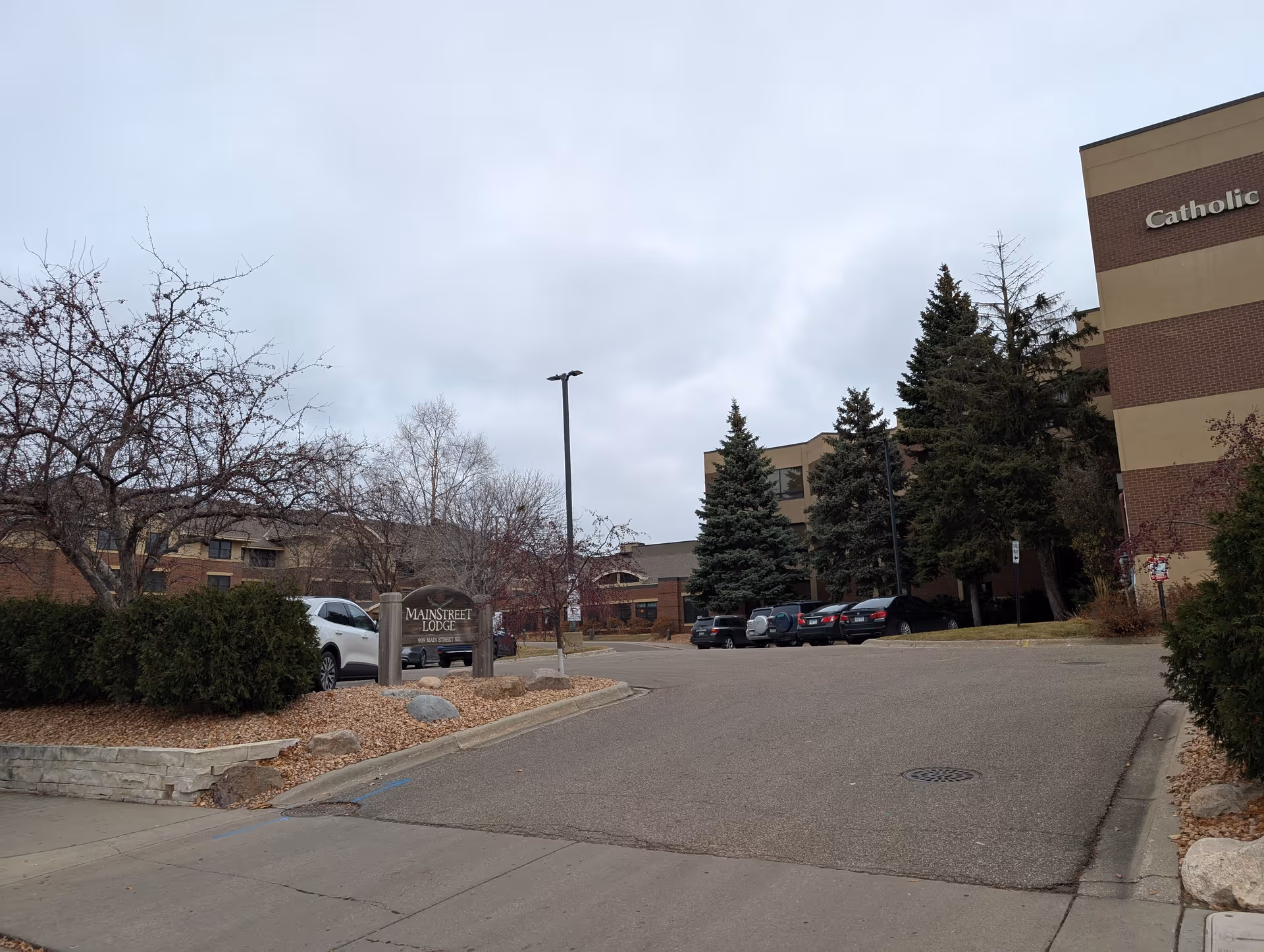 Entrance and parking area of Mainstreet Lodge with its sign, parked cars, trees, and a cloudy sky.