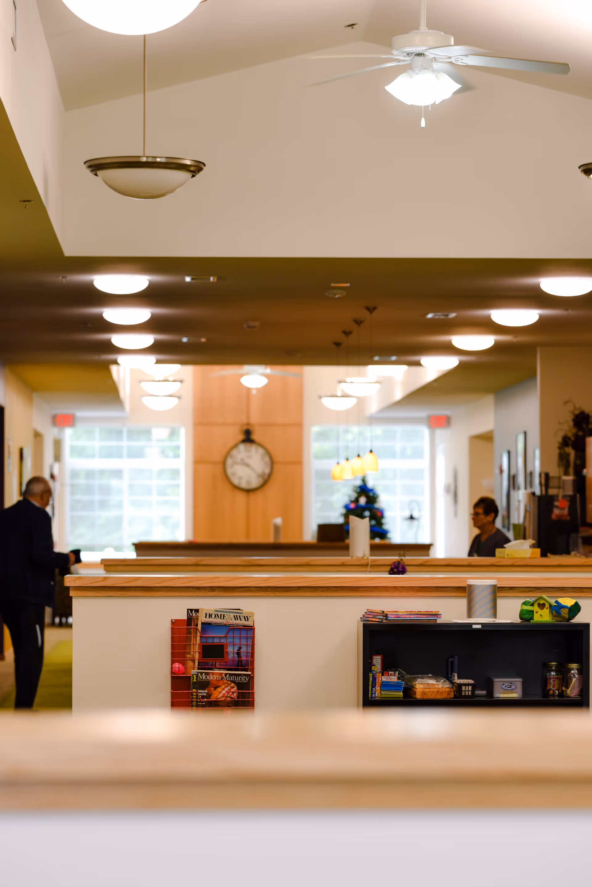 Interior view of a senior living facility hallway with a clock on the far wall, ceiling lights, and a ceiling fan. There are two people visible in the background, one walking and one seated. A small shelf with magazines and snacks is in the foreground.
