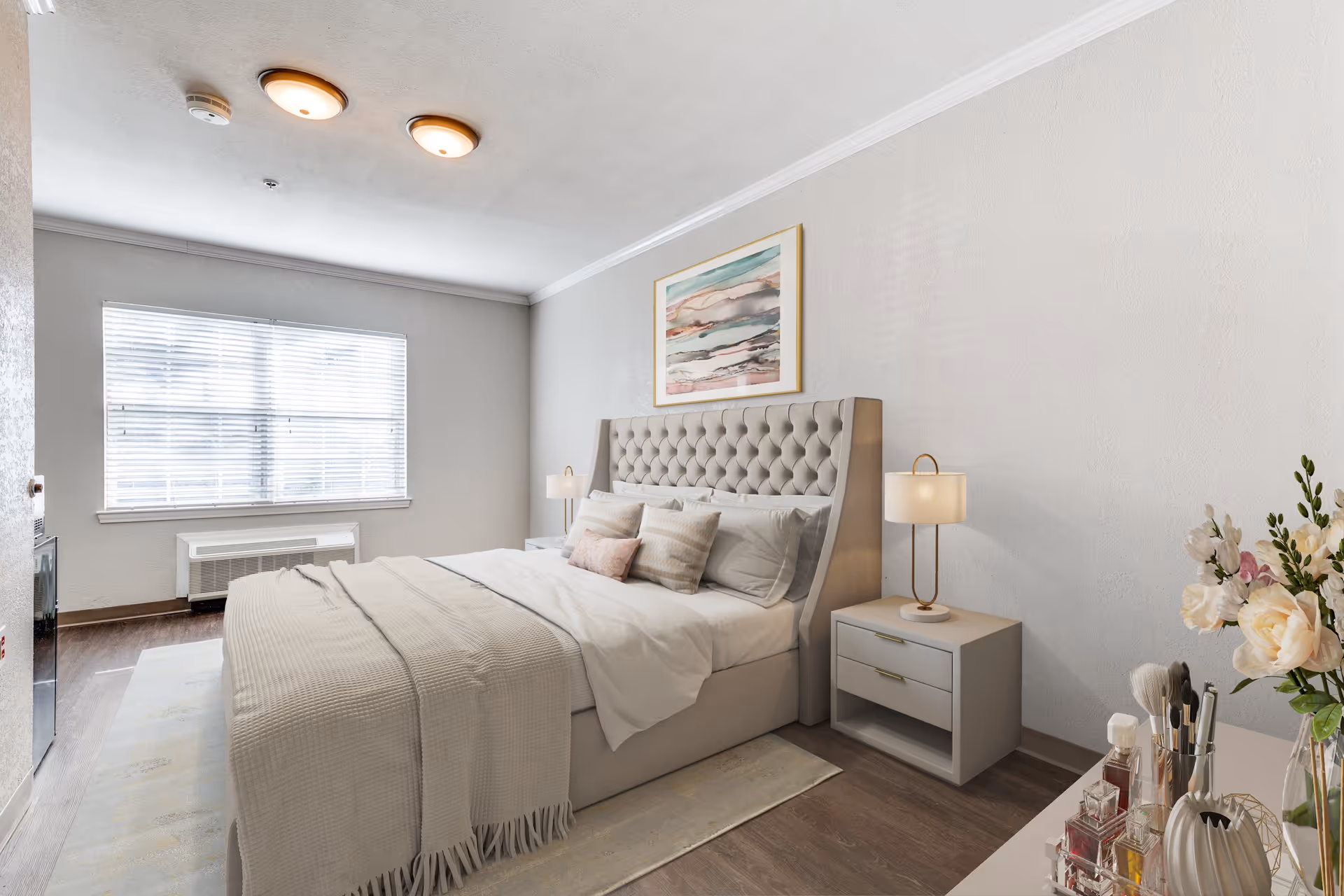 A bright and neatly arranged bedroom with a large bed featuring a tufted headboard, white and beige bedding, and multiple pillows. There are two bedside tables with lamps on either side of the bed. A large window with blinds allows natural light to fill the room. A framed abstract painting hangs above the bed, and a dresser with flowers and various personal items is visible in the foreground.
