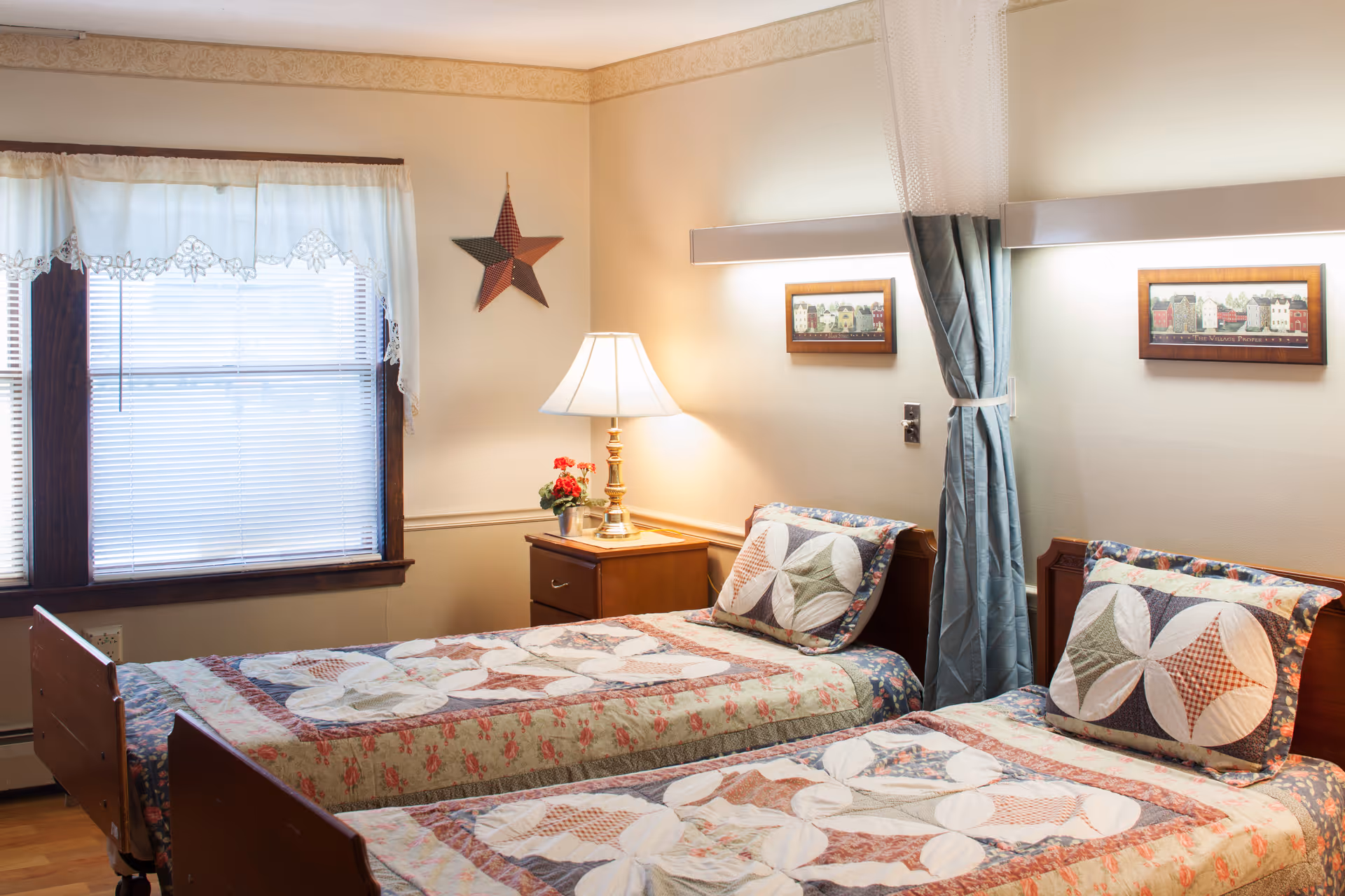 A cozy bedroom with two single beds side by side, each with a patterned quilt and matching pillow. A wooden nightstand with a lamp and a small flower pot is placed between the beds. A window with white lace curtains lets in natural light, and a decorative star hangs on the wall above the nightstand. Two framed pictures are mounted on the wall above the beds, and a privacy curtain is partially drawn between the beds.
