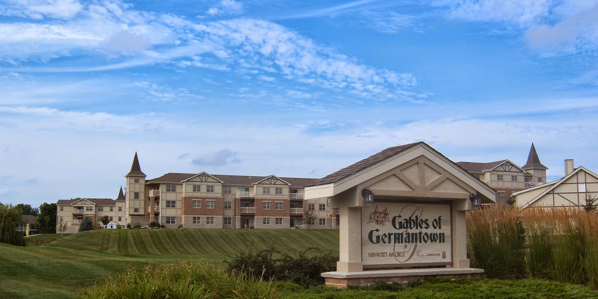 Exterior view of The Gables of Germantown senior living facility showing a large multi-story building with beige and brick facade, surrounded by well-maintained green lawns and landscaping under a partly cloudy blue sky. A prominent sign in the foreground displays the facility's name.