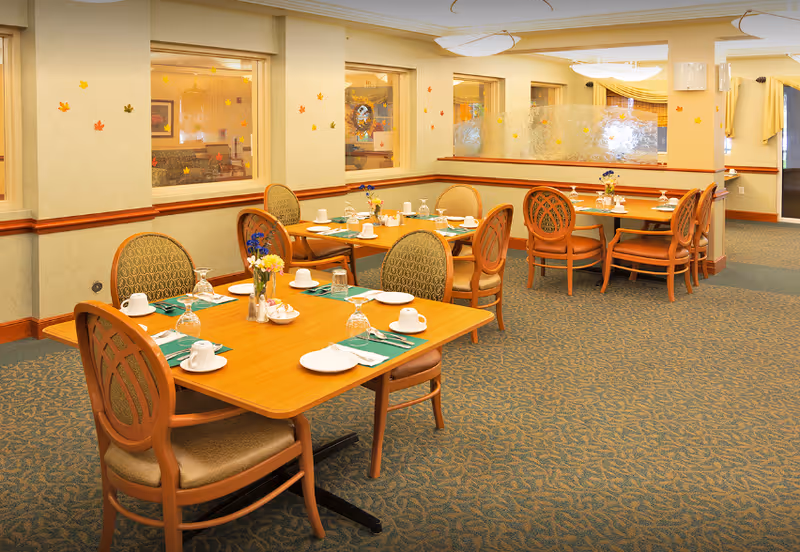 A dining room in a senior living facility with several wooden tables set with white plates, cups, glasses, and silverware. Each table has a small vase with flowers. The room has patterned carpet, beige walls with a wooden trim, and windows decorated with small colorful leaf decals. There are upholstered chairs around the tables and soft lighting from ceiling fixtures.
