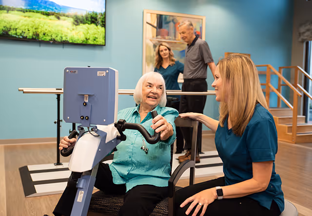An elderly woman using a seated exercise machine in a rehabilitation or therapy room, smiling and interacting with a female caregiver or therapist. In the background, a man and woman are walking with support rails. The room has light blue walls, a large TV screen displaying a nature scene, and wooden stairs with handrails.