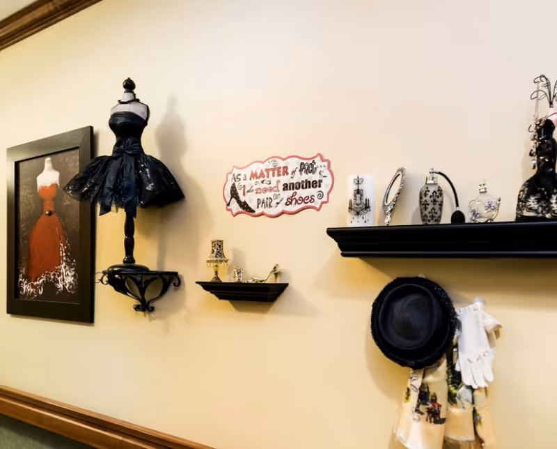 Wall decorated with a framed painting of a red dress, a black dress form with a black tutu, two black shelves holding decorative perfume bottles and jewelry, a sign that reads 'As a matter of fact I do need another pair of shoes', and a black hat with white gloves hanging below the shelves.