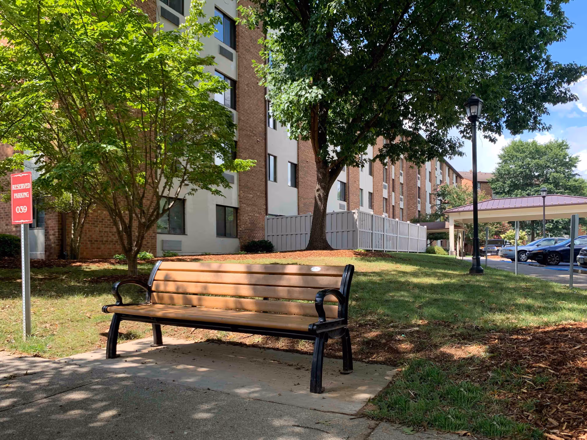A wooden bench with black metal armrests and legs sits on a concrete pad in a grassy outdoor area near a multi-story brick and beige building. There are several trees providing shade, a red sign indicating reserved parking spot 039, a lamppost, and a covered parking area with several parked cars in the background under a blue sky with some clouds.