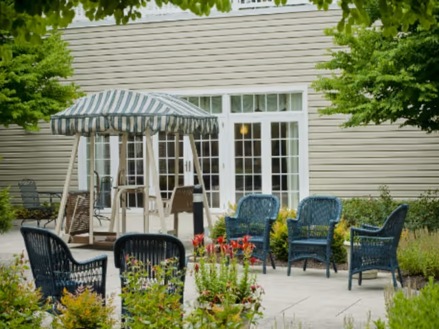 A landscaped outdoor patio with wicker chairs arranged around planters and a canopy-covered swing in front of glass double doors of a building.