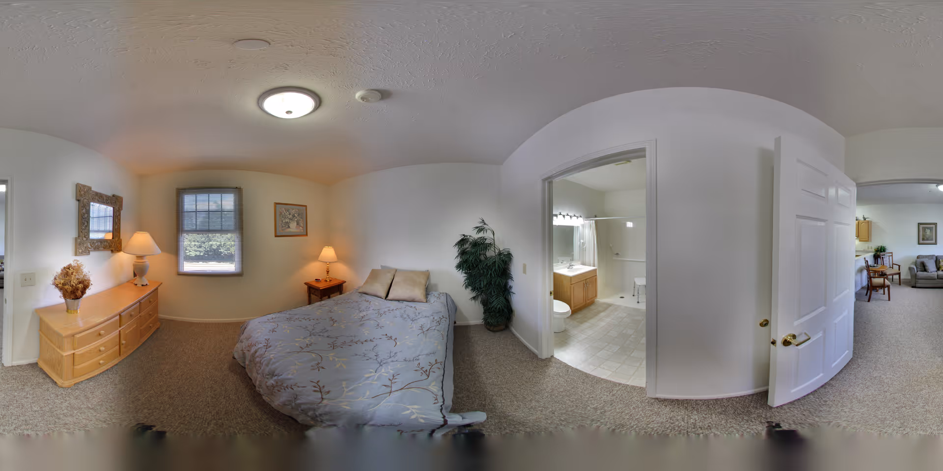 A cozy bedroom with a bed covered in a floral patterned comforter, two pillows, a wooden dresser with a lamp and a vase of dried flowers, a window with blinds, a framed picture on the wall, and a nightstand with a lamp. To the right, an open door leads to a bathroom with a vanity, sink, toilet, and a shower with a curtain. Another open door leads to a living area with chairs and a sofa.