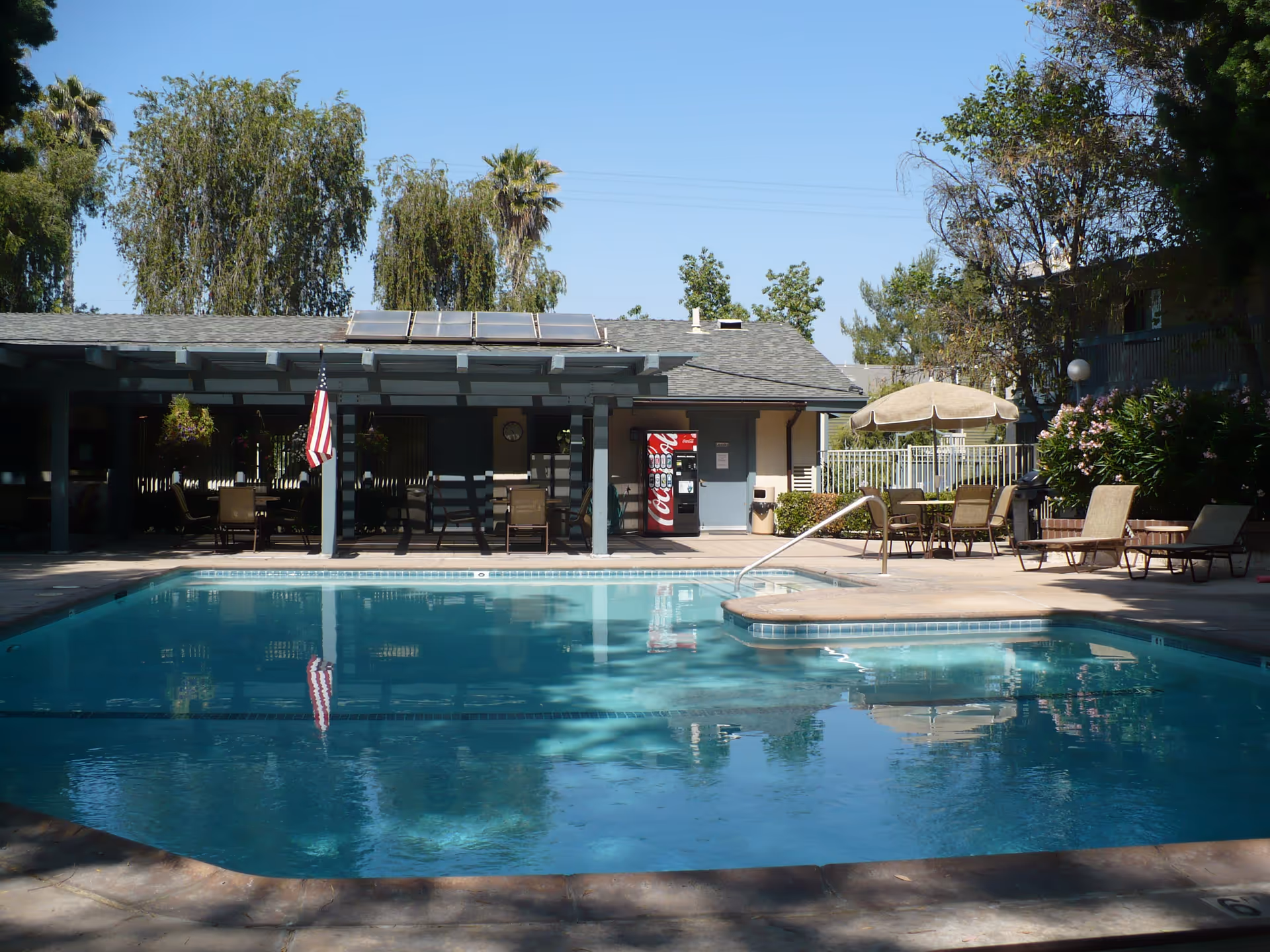 Outdoor swimming pool with lounge chairs, tables, an American flag and a covered patio area with a vending machine in the background.