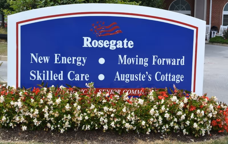 A blue and white sign for Rosegate senior living facility with the phrases 'New Energy', 'Moving Forward', 'Skilled Care', and 'Auguste's Cottage'. The sign is surrounded by red and white flowers and greenery, with part of a building and driveway visible in the background.