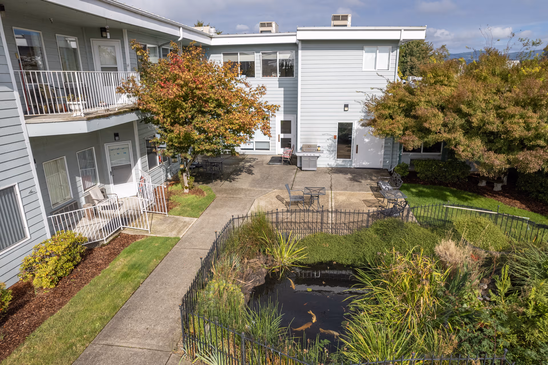 Courtyard of a two-story assisted living building with a koi pond, patio seating, trees, and walkways.