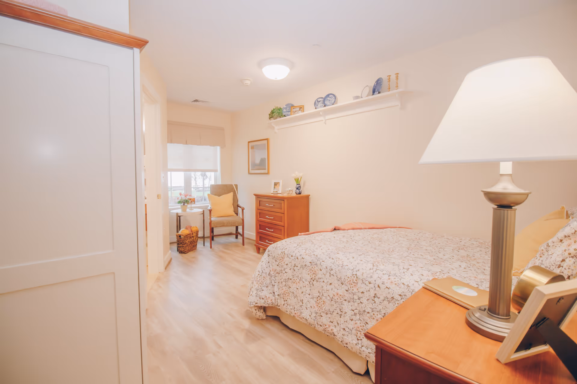 A cozy bedroom in a senior living facility featuring a bed with floral bedding, a wooden nightstand with a lamp and picture frames, a wooden dresser, a chair with a yellow cushion, a small round table with a plant, and decorative plates and items on a wall shelf. The room has light-colored walls and wood flooring with a window letting in natural light.