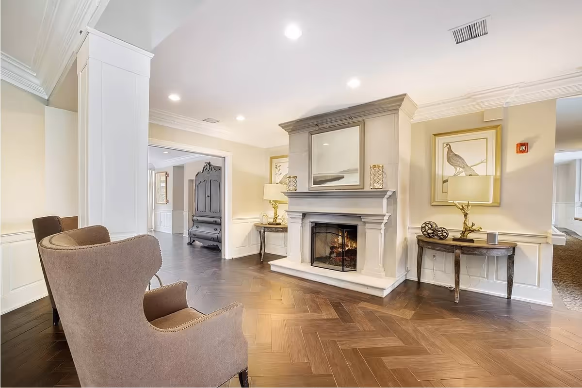 A cozy senior living common area featuring a lit fireplace with a large framed picture above it, two side tables with lamps and decorative items, a pair of upholstered armchairs, and wooden herringbone flooring. The walls are painted light beige with white wainscoting, and there is an open doorway leading to another room with a dark wooden cabinet.