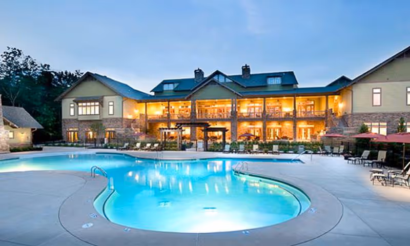 Outdoor view of a senior living facility at dusk featuring a large, illuminated swimming pool surrounded by lounge chairs and umbrellas. The building in the background has multiple windows with warm lights glowing inside, and the sky is clear with trees visible behind the building.