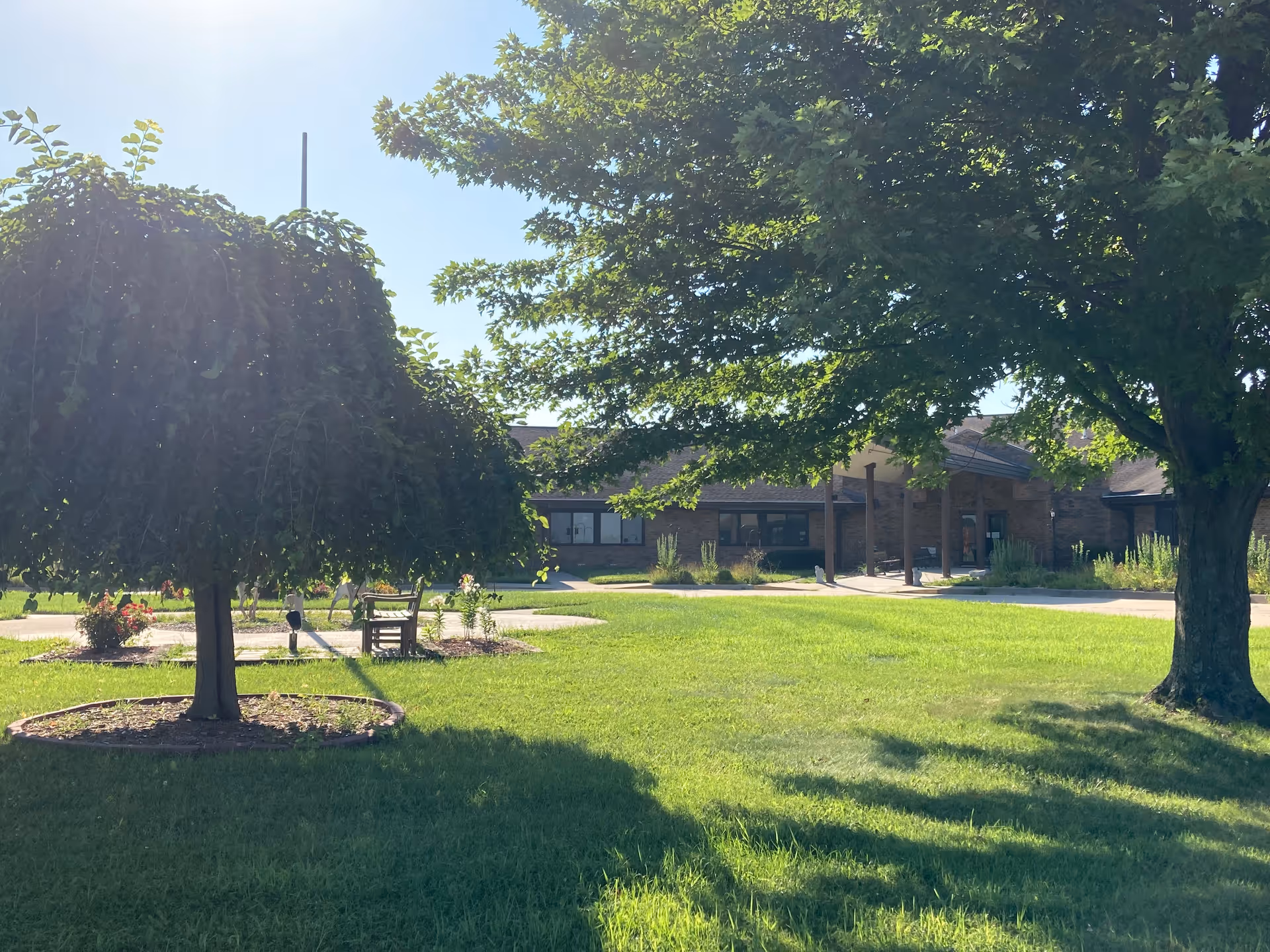 Grassy front lawn with trees and a bench in front of the single-story brick entrance of a senior living facility.