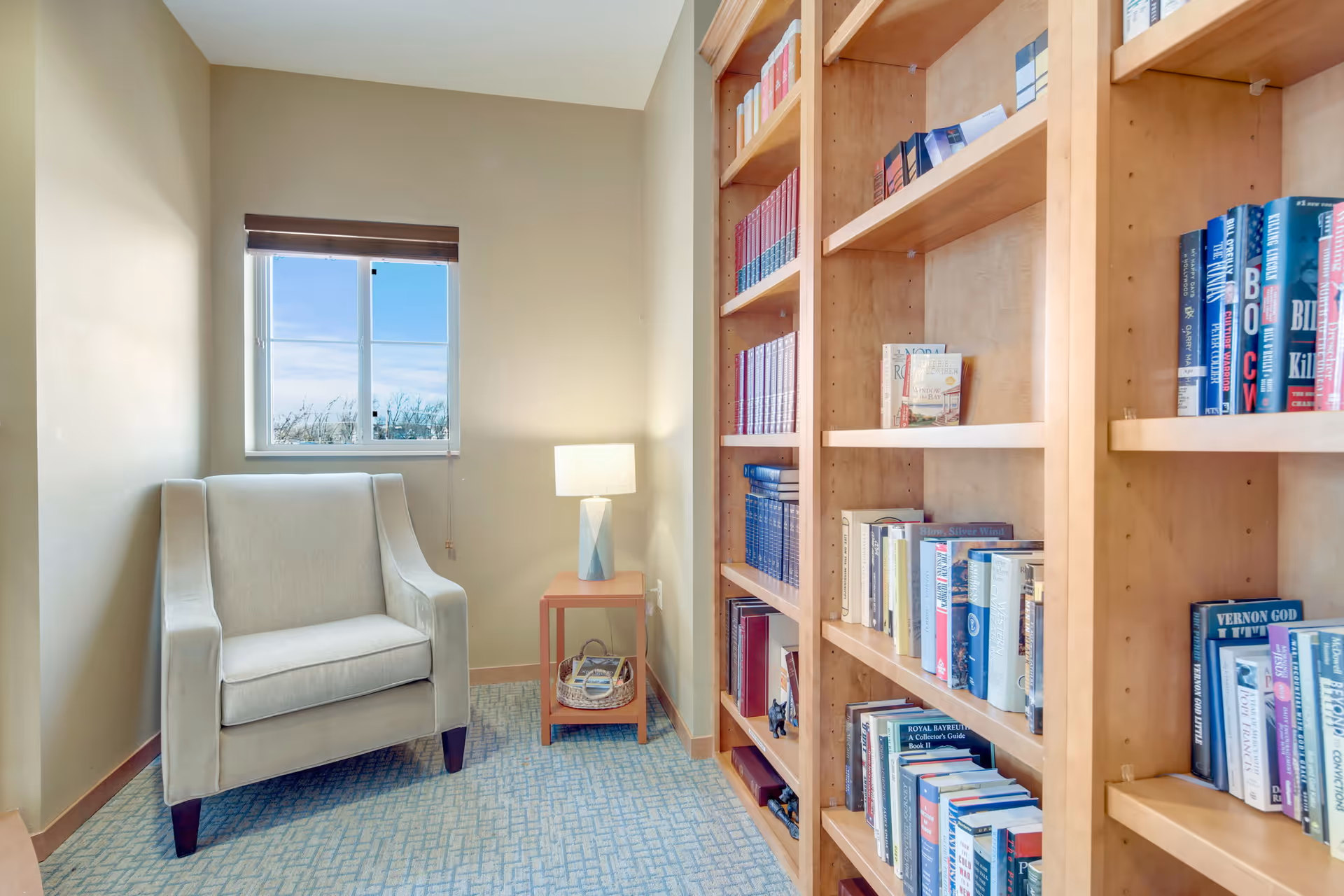 A cozy reading nook featuring a light gray armchair next to a small wooden side table with a lamp. To the right, there is a tall wooden bookshelf filled with various books. A window with a brown blind lets in natural light, illuminating the carpeted floor and beige walls.