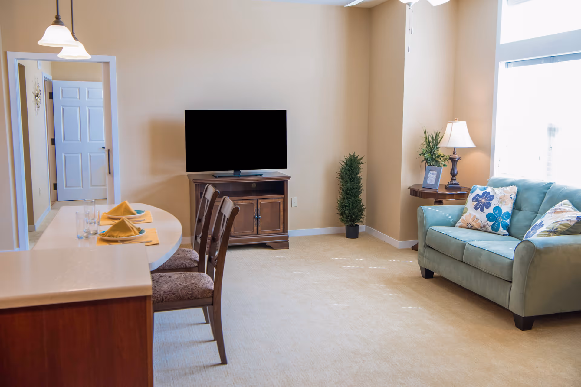 A bright living room area with a teal sofa adorned with floral pillows, a wooden side table with a lamp and a small plant, a flat-screen TV on a wooden stand, and a dining counter with two chairs and place settings with yellow napkins and glasses. The room has beige walls and carpet, with a doorway leading to another room.