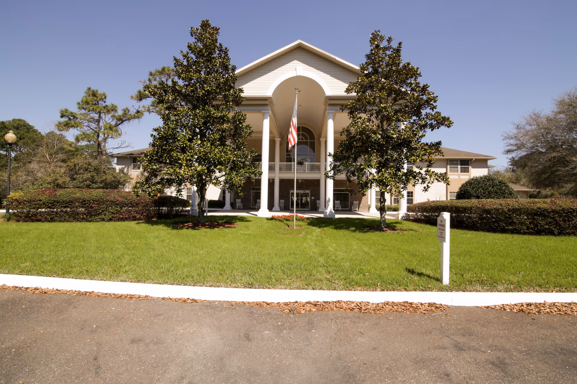 Front exterior view of a senior living facility building with a large covered entrance supported by white columns, an American flag on a flagpole in the center, green lawn, trees, and clear blue sky.