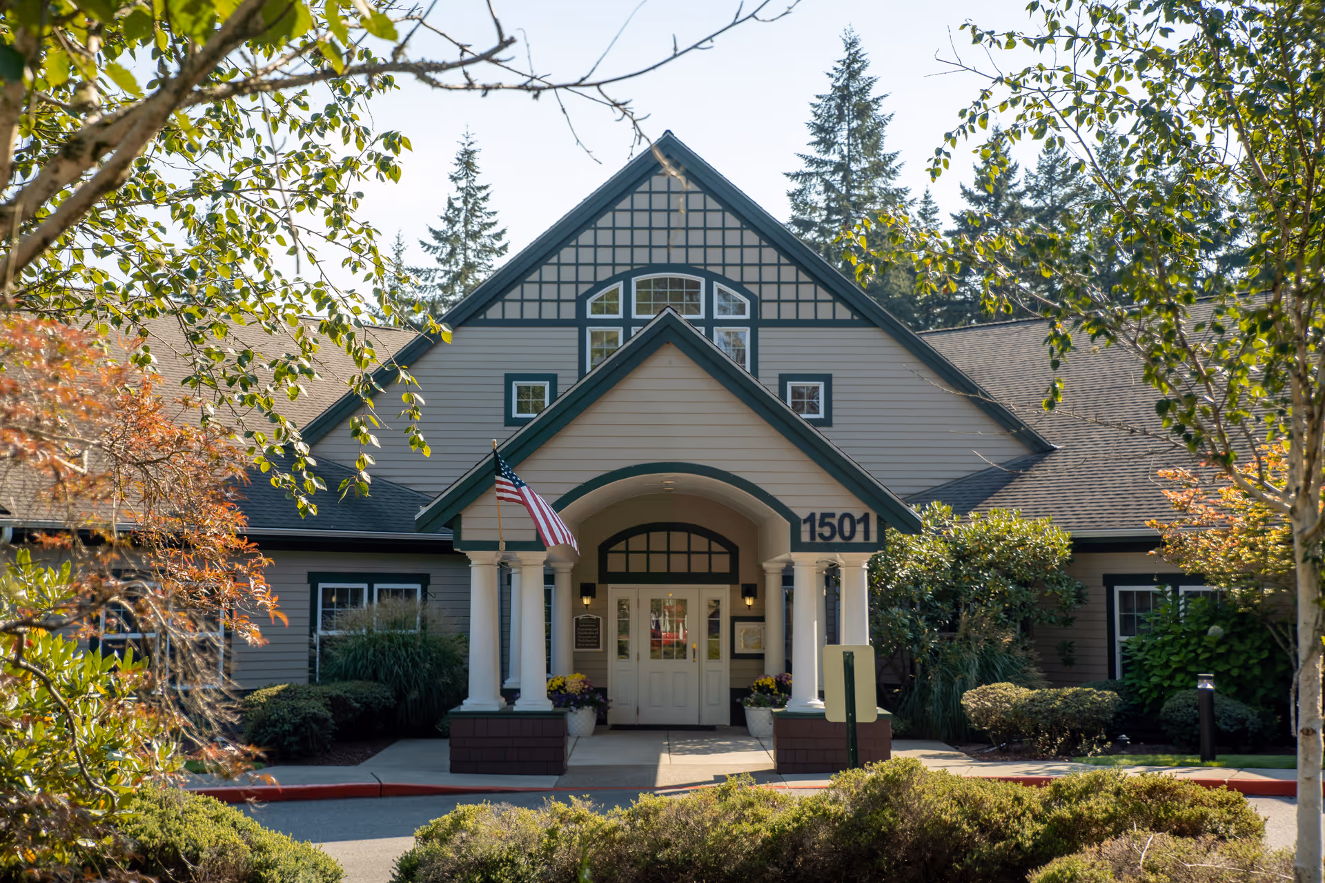 Front exterior view of a senior living facility named The Ridge an Encore Community, featuring a peaked roof with decorative trim, white columns at the entrance, an American flag, and surrounded by trees and shrubs.