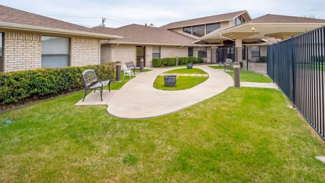 Outdoor courtyard area at Rowlett Health & Rehabilitation Center featuring a curved concrete walkway, green grass, benches along the path, shrubs along the building, and a black metal fence on the right side.