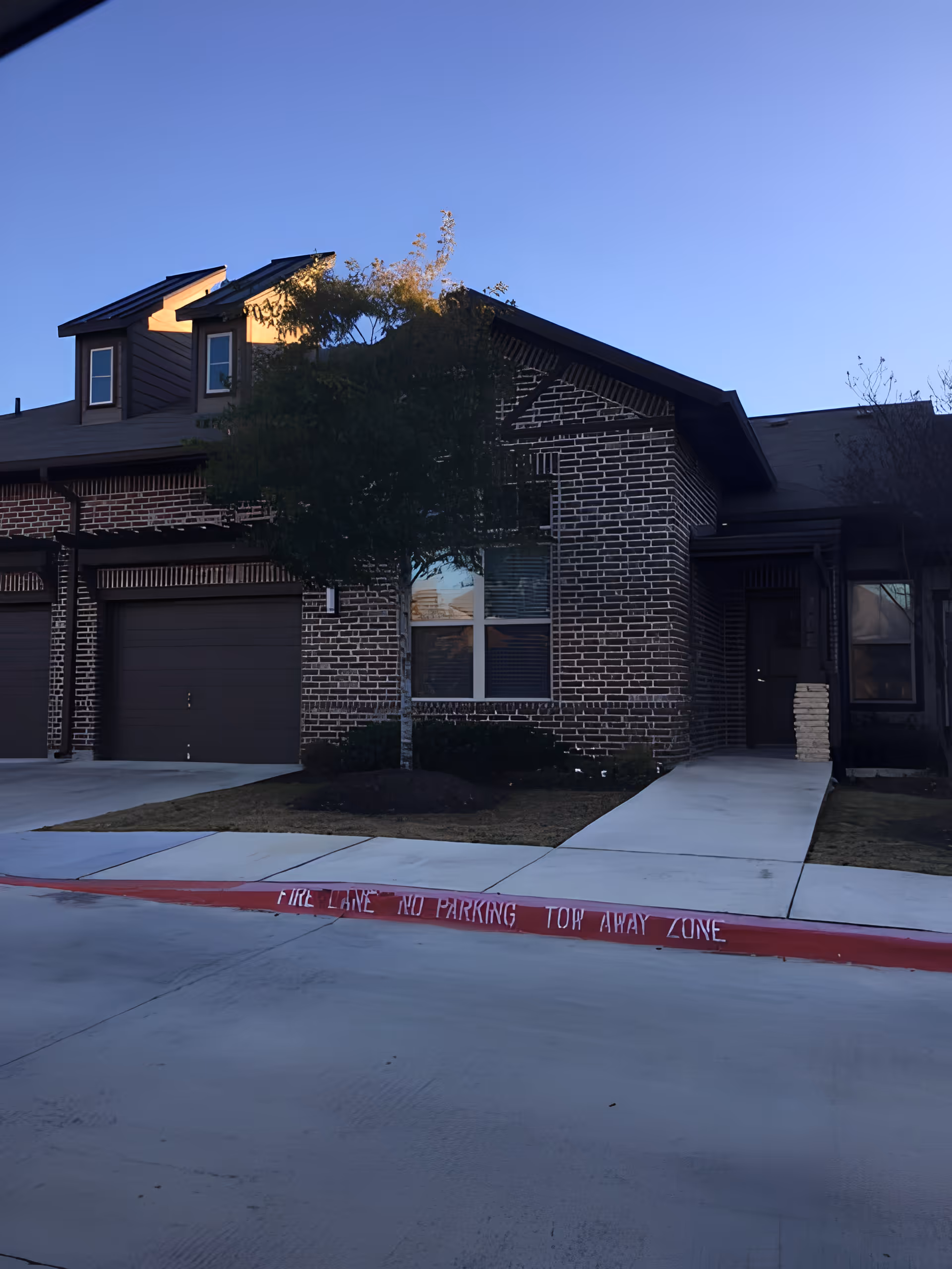 Brick townhouse front with two garage doors, a central window, small tree, walkway, and a red curb marked fire lane.