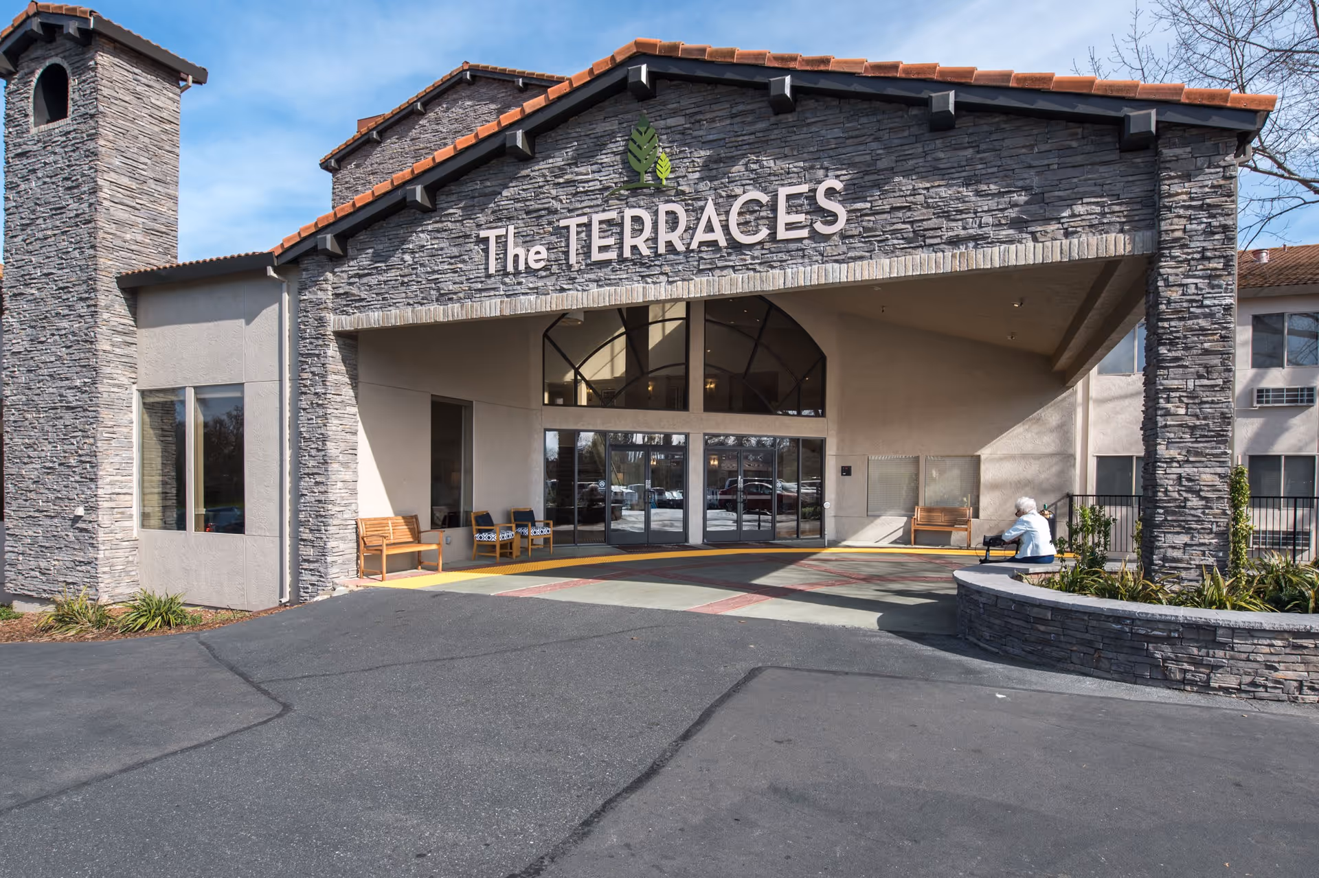 Exterior view of The Terraces senior living facility entrance with stone facade, benches, and a person sitting on a low stone wall near the entrance.