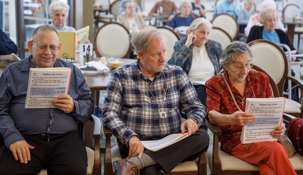 A group of elderly people sitting in a common area of a senior living facility, some holding and reading booklets. The room has rows of chairs and tables, with several other seniors visible in the background engaged in various activities.