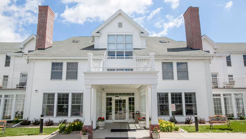 Front exterior view of a large white senior living facility building with multiple windows, two prominent brick chimneys, a covered entrance with columns, flower pots, benches, and a clear blue sky with some clouds.