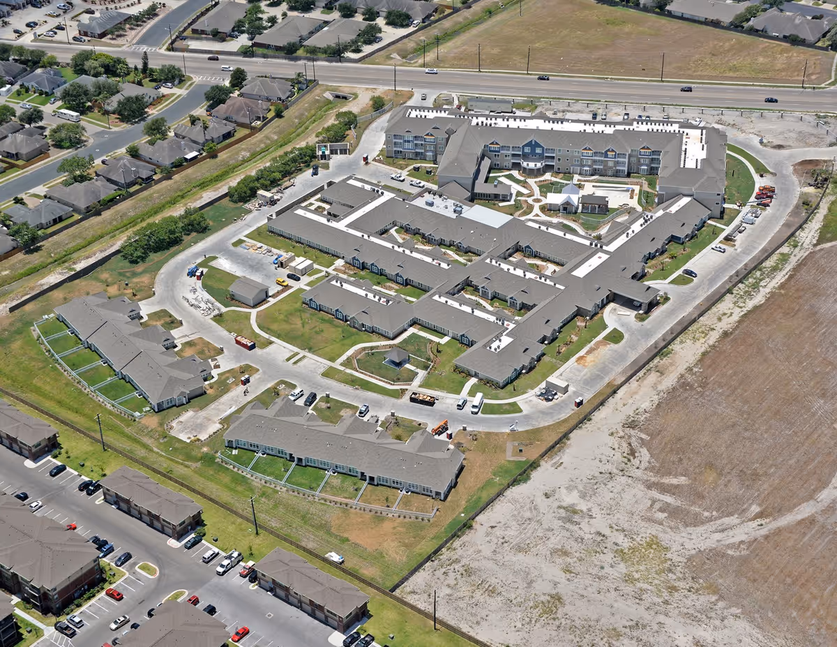 Aerial view of The Viera Senior Living facility showing multiple connected buildings with gray roofs, surrounding green lawns, parking areas, and nearby residential neighborhood. The facility is bordered by roads and open land.