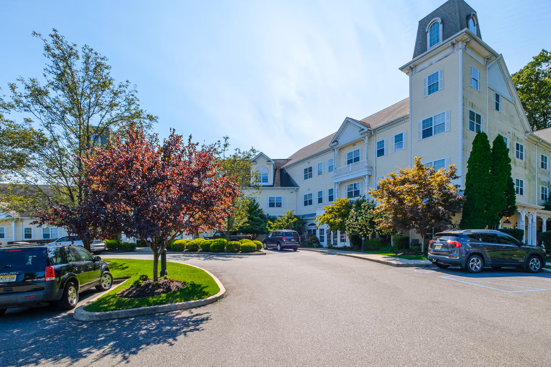 Exterior view of a large, multi-story senior living facility building with cream-colored siding and multiple windows. The building is surrounded by well-maintained landscaping including trees and bushes. Several cars are parked in the parking lot in front of the building under a clear blue sky.