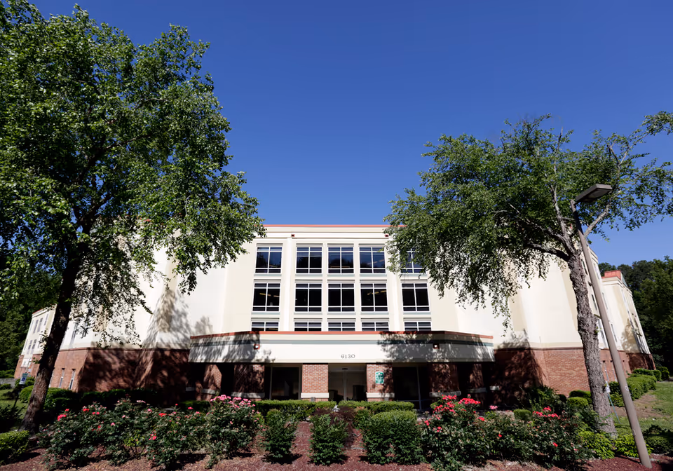 A multi-story senior living facility with a white facade, brick base, large windows, and landscaped entry under a clear blue sky.