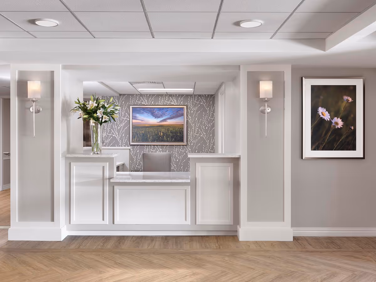 A clean and modern reception desk area with white paneling and a marble countertop. Behind the desk is a gray wall with a subtle white branch pattern, a framed landscape photograph of a field under a colorful sky, and a gray chair. On the left side of the desk is a vase with white lilies. The walls are painted light gray with two wall sconces and a framed photo of flowers on the right side. The floor has a light wood pattern.