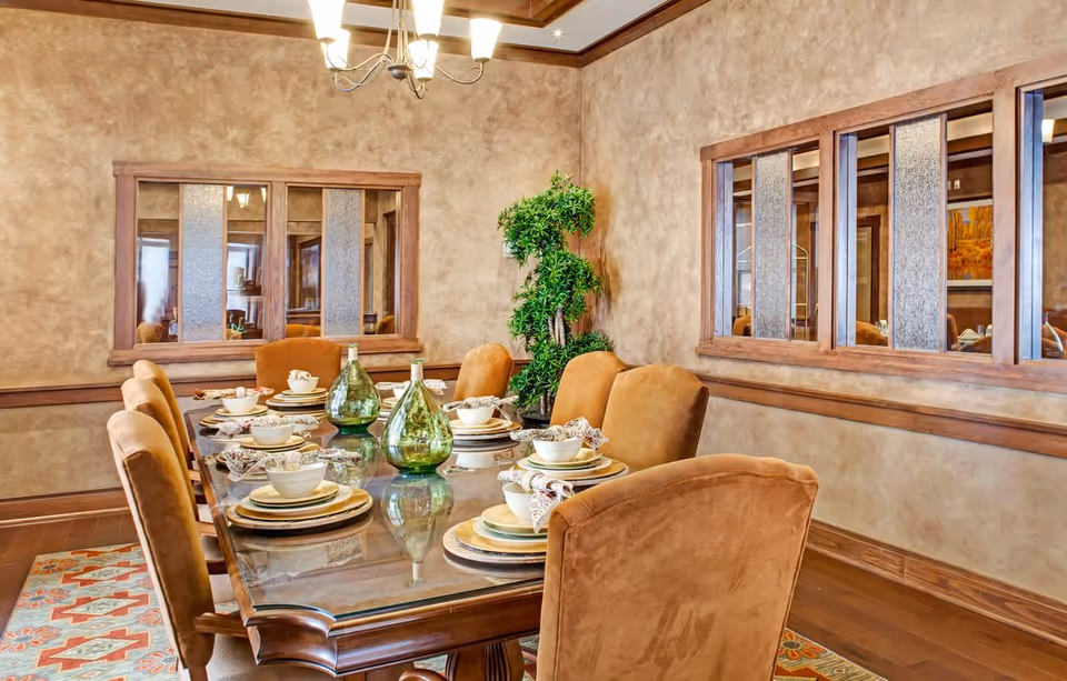 A dining room with a glass-top wooden table set for eight people. The table is arranged with plates, bowls, napkins, and two green decorative glass bottles. The room has beige textured walls, wooden trim, a chandelier overhead, and a leafy green plant in the corner. There are two large windows with wooden frames on the walls.
