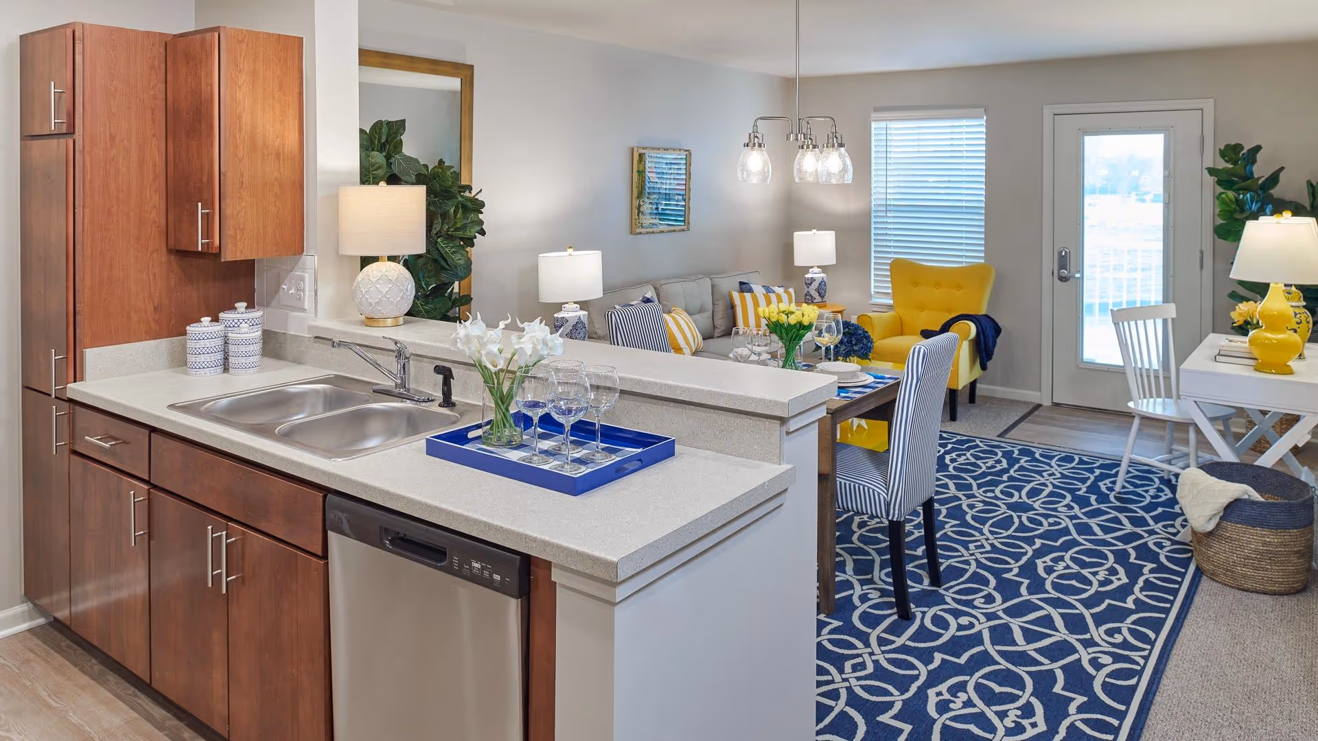 View of a modern open-concept kitchen and living area in a senior living facility. The kitchen features wooden cabinets, a double sink, and a dishwasher. A countertop with a tray holding wine glasses and flowers separates the kitchen from the living space. The living area includes a dining table set with plates and glasses, a yellow armchair, a sofa with striped and yellow pillows, a blue patterned rug, and a small desk with a lamp and plant near a glass door leading outside.