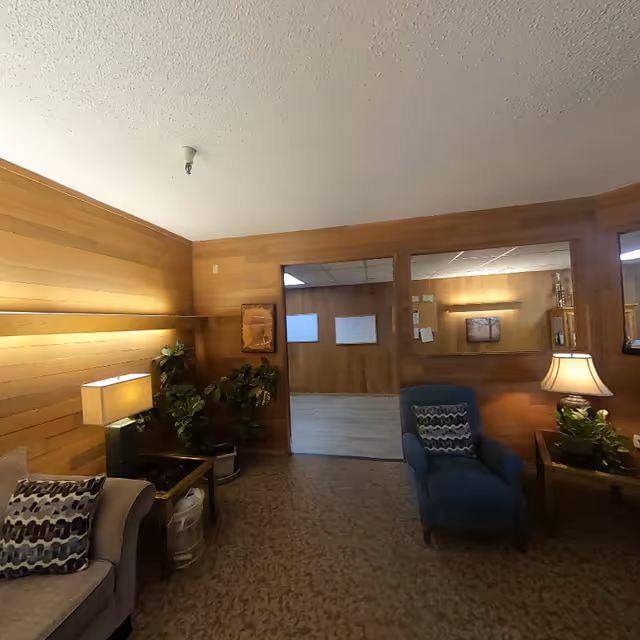 A cozy interior room with wood-paneled walls and carpeted floor. The room features a beige couch with a patterned pillow, a blue armchair with a matching pillow, two side tables with lamps, and several potted plants. There is an open doorway leading to another room with wood-paneled walls and a light-colored floor. The lighting is warm and inviting.